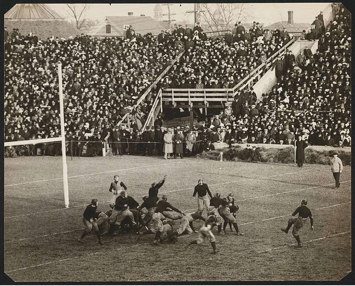 Crowds watch football game between Harvard and Dartmouth with players on the field as ball is kicked 