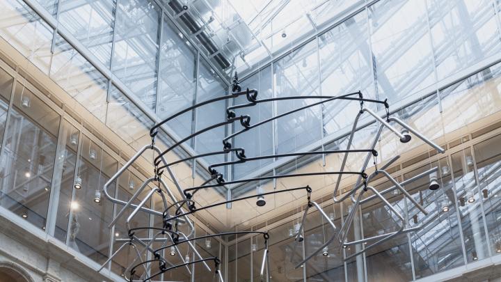A chrome-plated triangle dangles from the end of each painted steel bar, their weight carefully distributed to achieve equilibrium. The joints of the sculpture allow it to rotate freely in the air.