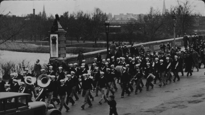 Band members wearing sailor hats march across a bridge to play at a football game