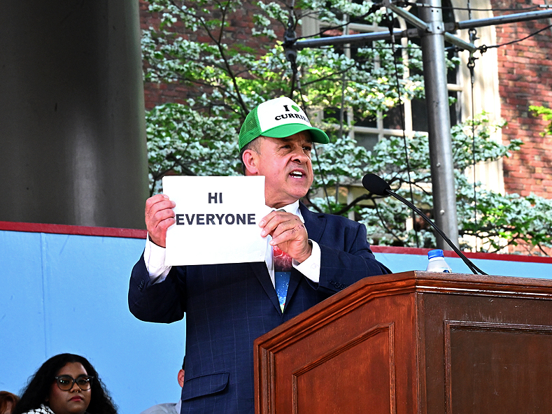 Man with "I love Currier" hat and a sign that reads "Hi Everyone" speaks at podium