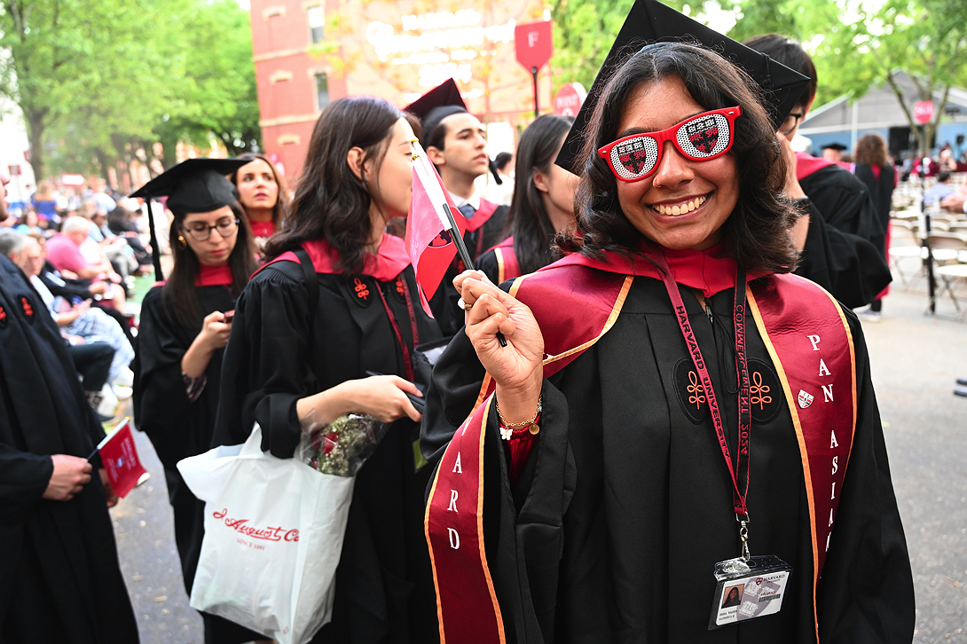 Student in black gown waves red flag and wears novelty Harvard sunglasses