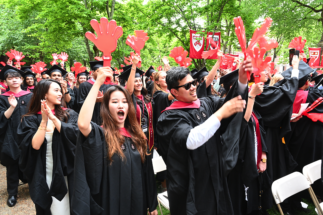 graduates wearing black gowns hold large plastic red clapping hands and cheer