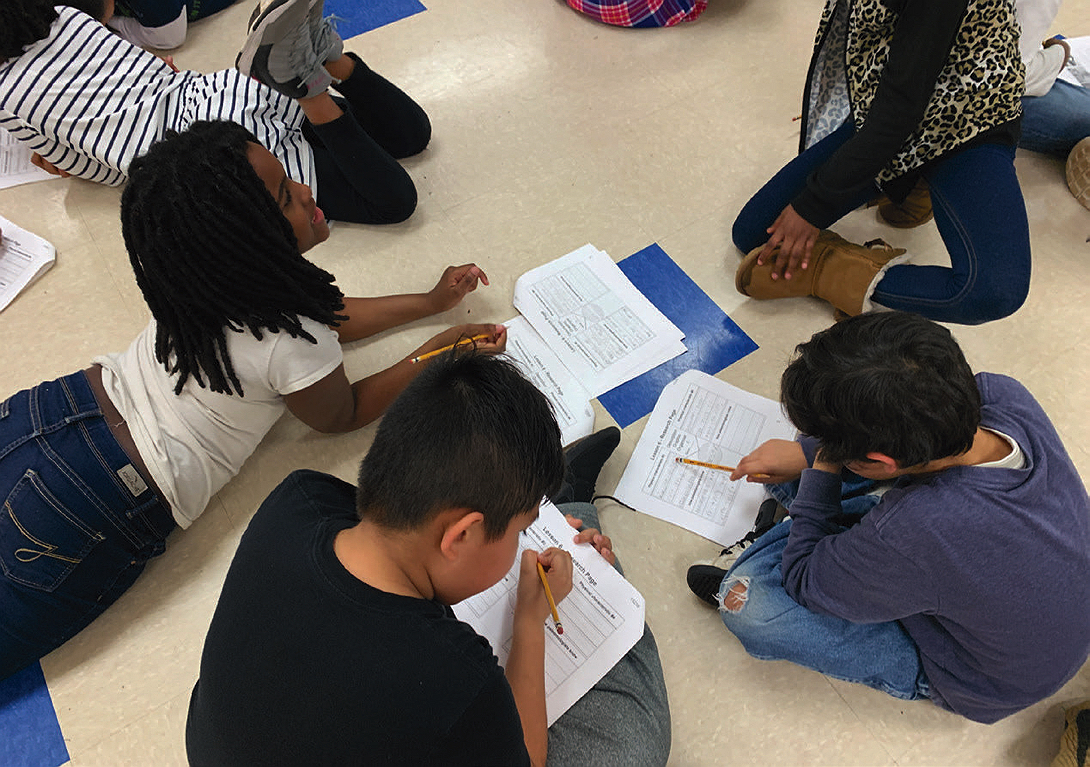 Students sit and lie on the floor in a group, collaborating on worksheets with pencils in hand.