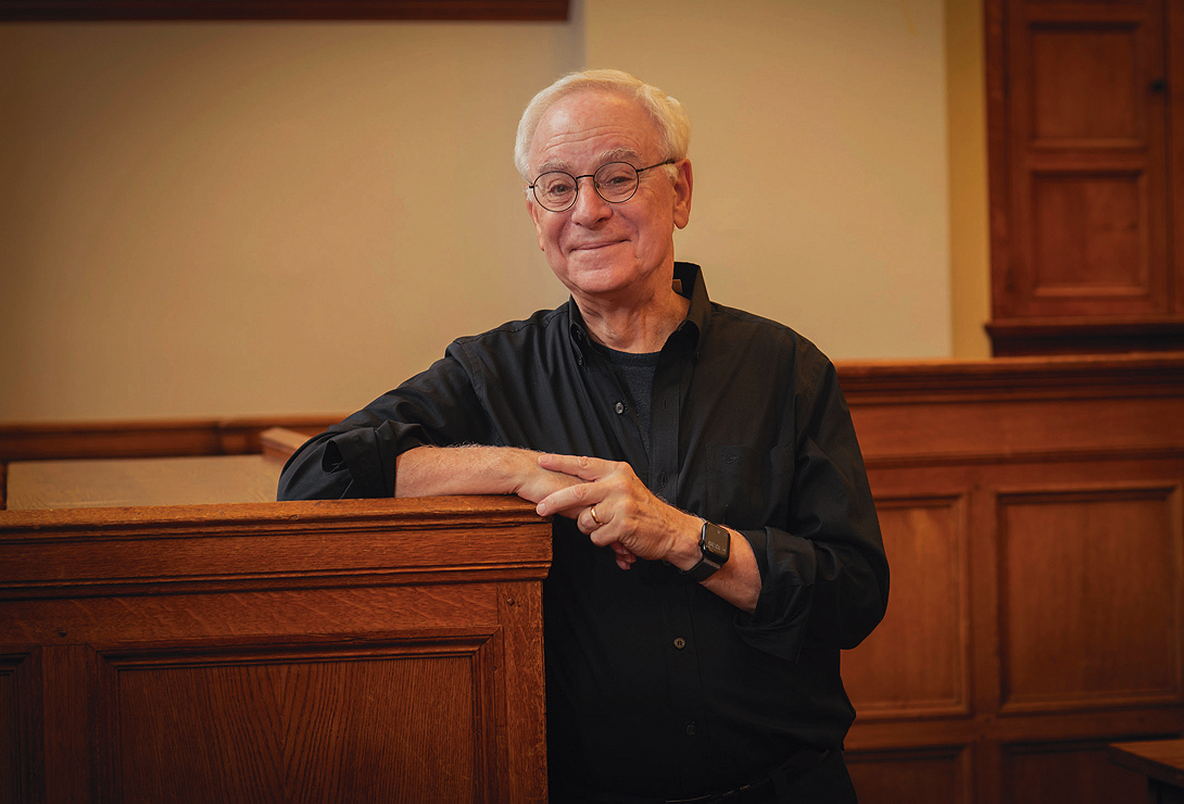 Robert C. Post, wearing a black shirt and glasses, leans casually against a wooden railing in a warmly lit room, smiling.