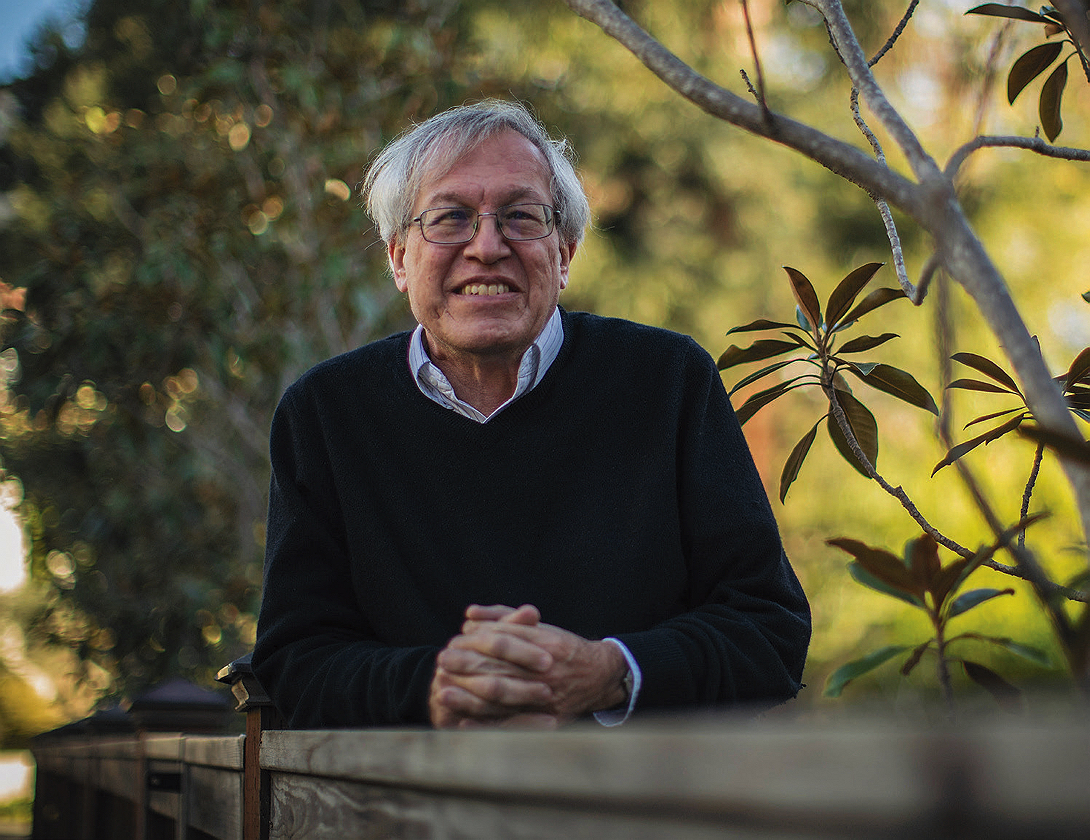 Erwin Chemerinsky, wearing glasses and a dark sweater, leans on a wooden railing outdoors, smiling with trees in the background.