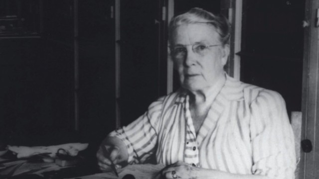 Photograph of a stout middle-aged woman seated at her desk, writing