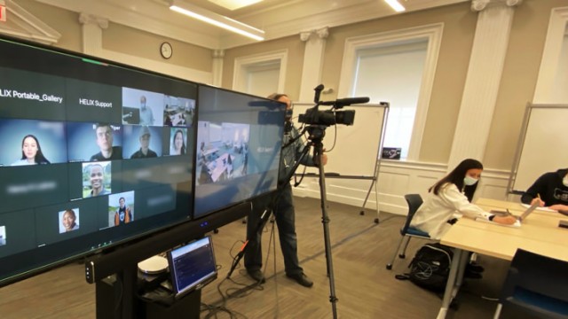 A hybrid classroom showing the large screens enabling a teacher to see all the students, remote or present, as he is followed by a camera operator.