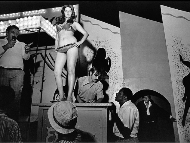 Photograph by Susan Meiselas/Magnum Photos “Bally call,” Essex Junction, Vermont, 1973. (The call, performed by a “talker,” is used to entice men into the show. ) “Watch it! Watch it right now! You’re gonna see burlesque, striptease, hootchie-kootchie, and daddy-o it’s all the way. The show starts right now—now’s the time to go.” —From <em>Carnival Strippers</em>