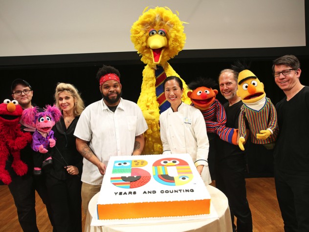 Two bakers present a large birthday cake, surrounded by Muppets and their puppeteers.