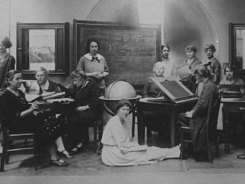 Several Harvard Computers and Radcliffe students pose for a photograph together, seated at desks and sitting on the floor