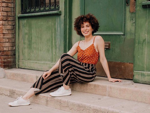 Adriana Colón, a casually dressed, smiling young woman with curly brown hair, sits in a doorway.