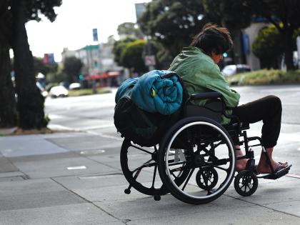 A man in a wheelchair with a rolled-up sleeping bag strapped to the back