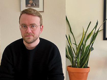 Richard Beck sitting indoors beside a potted plant, hands clasped, looking at the camera.