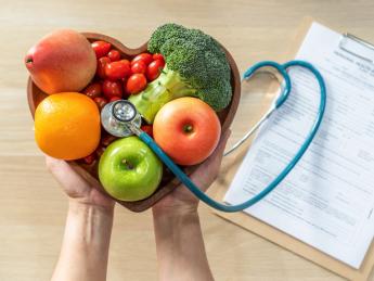 Hands holding a heart-shaped bowl of fruits and vegetables with a stethoscope, clipboard with medical records in the background.