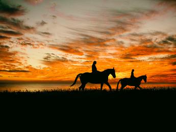 Photograph of two riders on horseback crossing a plain, silhouetted against the setting sun