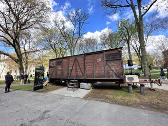 A red-brown train car with boarded-up windows, harsh metal siding, and faded industrial German lettering is on display in Harvard Yard.