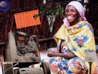A grinning woman in traditional Nigerian dress sits cross-legged on the floor surrounded by modern devices, including a power strip, a land-line telephone, and a desktop computer displaying on its screen a duplicate image of the entire montage.