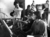College students, one with a trump, another with an upright bass, gather around a piano with Dizzy Gillespie, who plays his trumpet while a student pianist also plays 