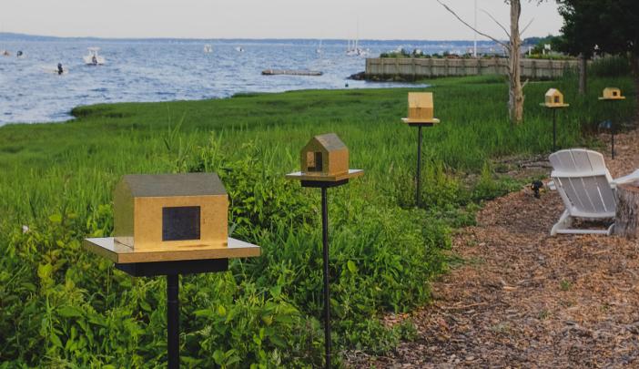 five small sculptures of houses made of brass, suspended on posts and placed on the grassy shore overlooking Duxbury Bay