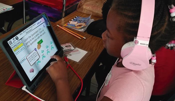 A young girl with pink headphones uses a tablet for an educational reading game at a classroom desk.