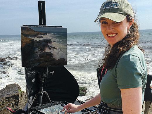 Hudson in a baseball cap with her easel and paints at a rocky seaside