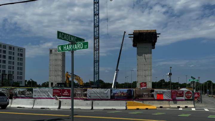Construction site at the intersection of Academic Way and North Harvard Street. Two tall, unfinished concrete structures stand behind a fenced area with various signs and banners.