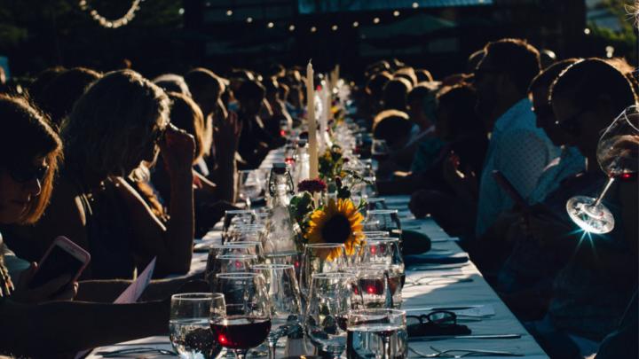 People gathered at a long, outdoor dining table set with wine glasses, candles, and flowers. 