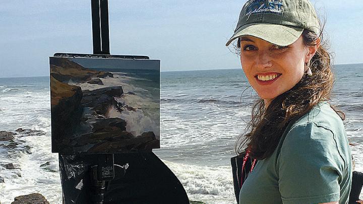 Hudson in a baseball cap with her easel and paints at a rocky seaside