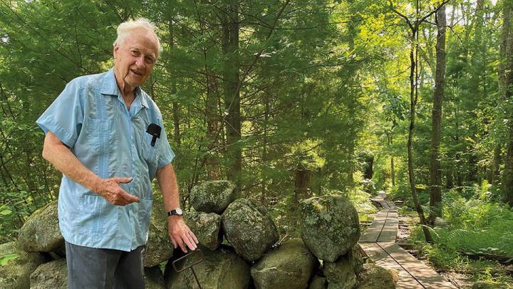 A white haired professor emeritus standing by a stone wall in the woods explaining its origins 