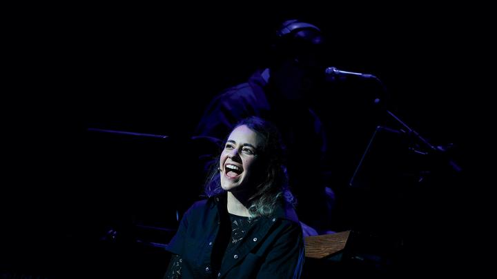 Madeline Benson singing at the piano, her face lit up on an otherwise dark stage