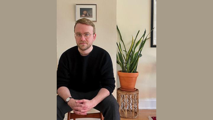 Richard Beck sitting indoors beside a potted plant, hands clasped, looking at the camera.