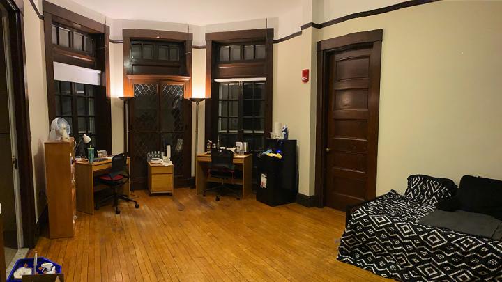 A large dorm room with desk, bed, lamps, and quarantine necessities, including bottled water, masks, and sanitizing supplies.