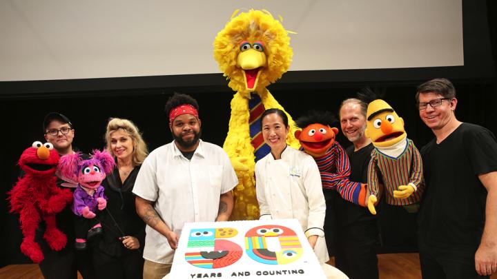 Two bakers present a large birthday cake, surrounded by Muppets and their puppeteers.